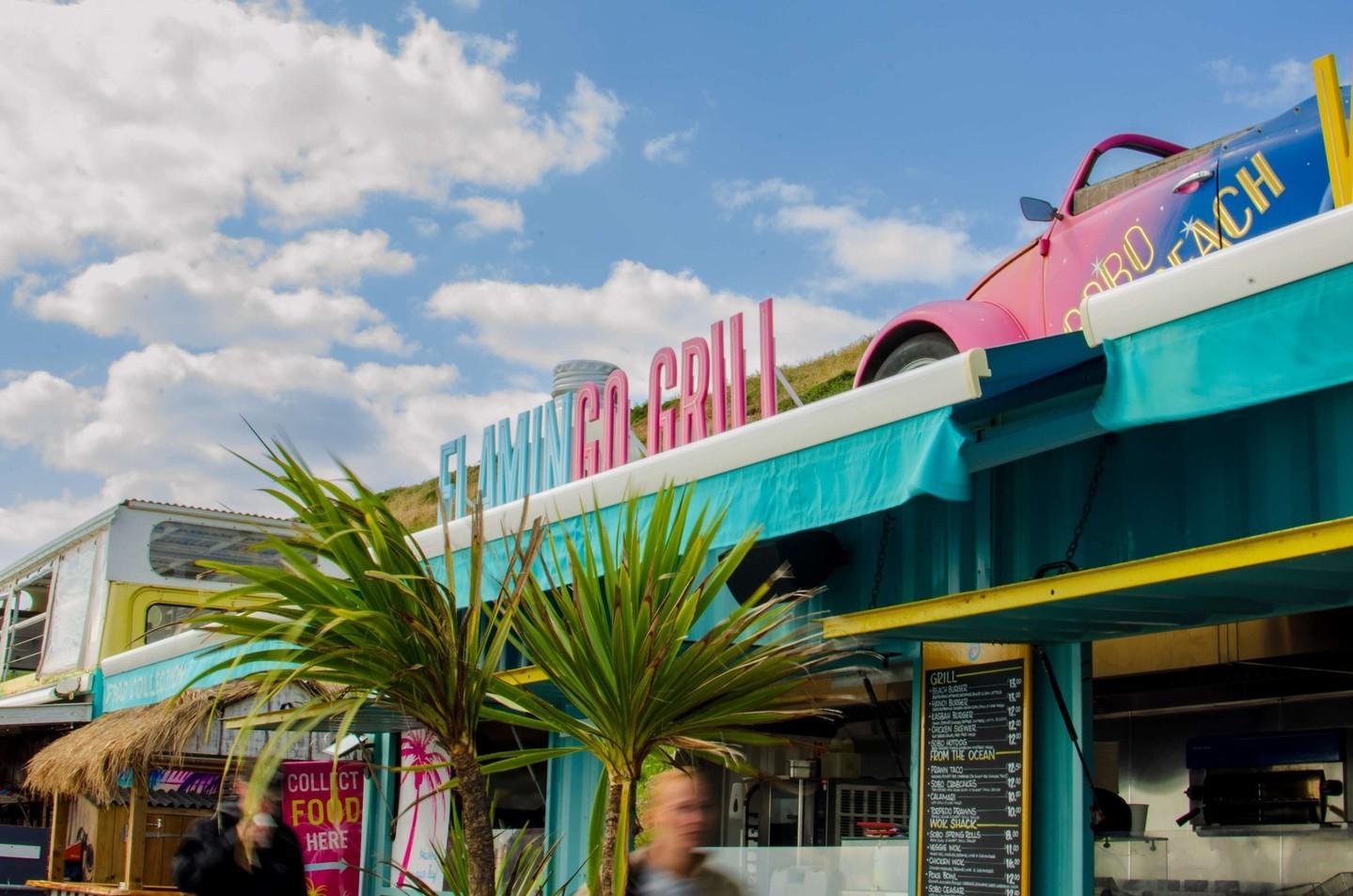 The pink Flamingo Grill neon sign and SOBO Beach’s painted Beetle above the teal food kiosks, under a blue sky