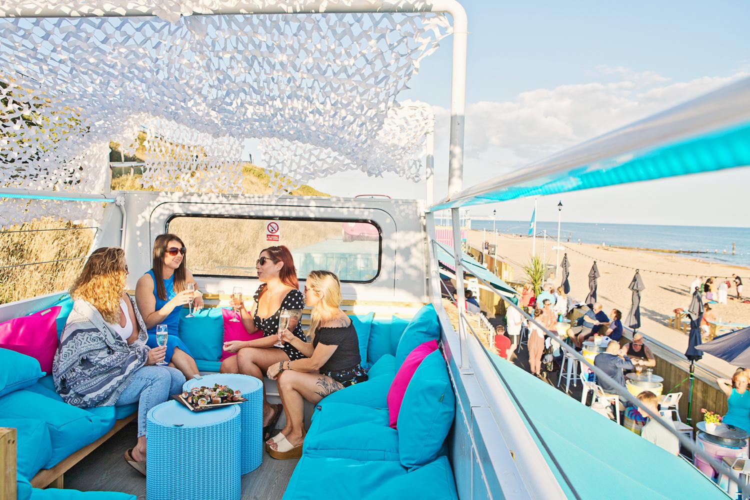 Friends enjoying drinks together on the sunny upper deck of SOBO Beach’s bus, with the beach and sea beyond