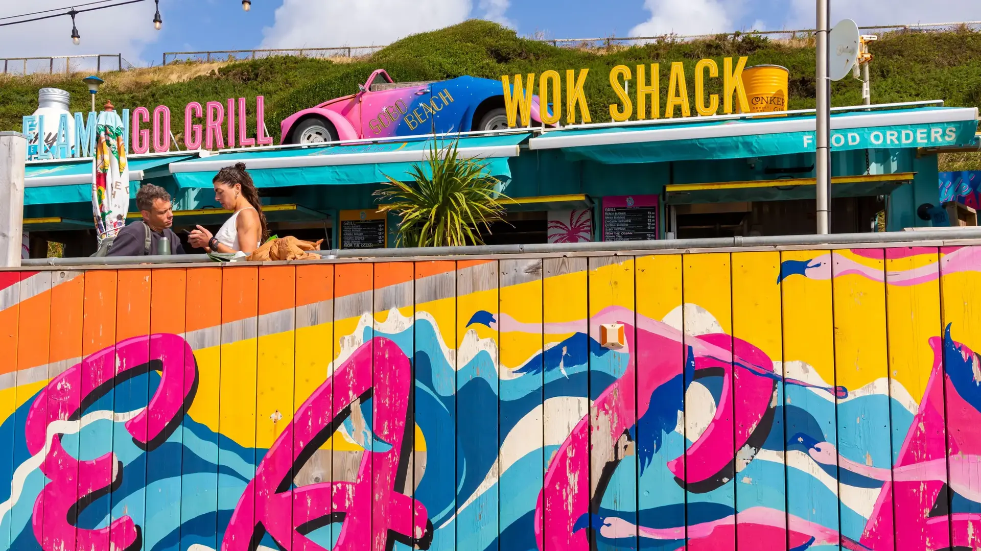 Two people chatting beside SOBO Beach’s hand-painted flamingo and wave mural, below the Flamingo Grill and Wok Shack signs